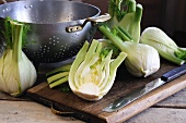 Fennel bulbs, whole and halved, on a chopping board with a knife and colander