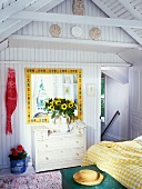 Sonniger Schlafraum mit farbenfroh gerahmtem Spiegel, einem gelb-weissen Bettbezug und Kinderhandabdrücke auf einer Konsole unter dem Dachgiebel
