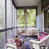 Verglaste, sonnige Veranda mit Hollywoodschaukel und Korbmöbeln; darauf Dekokissen mit Karo- und mit floralem Muster