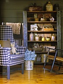 Vintage-style wooden shelving holding collection of antiques next to armchair with blue and white gingham upholstery and rocking chair