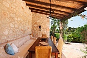 Tisch mit Stühlen und Sitzbank auf überdachter Terrasse vor Natursteinwand und Gartenblick