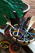 Wooden salad servers in bowl and colourful, African-style dishes on floor next to potted plant