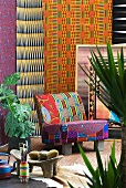 African Style - Farbig gemusterter Sessel vor bunten Tüchern zwischen tropischen Pflanzen im Topf