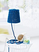 Einfache Tischlampe, aufgepeppt mit grob gestricktem, blauem Lampenschirm und dekorativ drapiertem Garnende