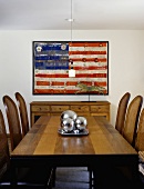 Dark wood dining table and rush-back chairs in front of painting of American flag on wall