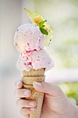 A hand holding a strawberry and yoghurt ice cream cone with almond brittle