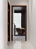 View of free-standing designer bathtub behind glass wall along glossy white corridor though open bathroom door