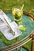A mojito next to an ice cube tray and bar tongs on a tea trolley