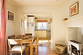 Dining area with simple wooden furniture next to antique stove in masonry fireplace and view into white country-house kitchen