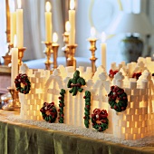 An illuminated house made from sugar cubes, on a table in front of candle holders with lit candles