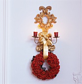 Gilt wall bracket with lit candles and wreath of red berries on white wooden wall