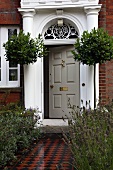Bay trees flanking stately entrance of English country house with brick facade and mosaic path edged by beds of lavender