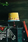 Honey yellow lampshade decorated with applied flowers and pompom trim next to colourful crochet blanket and fake zebra-skin blanket
