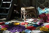 In verschiedenen Farben gehäkelte Blumenmotive und buntes Dekokissen vor Flechthocker auf Holzboden