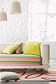 Sofa with striped cover and scatter cushions on red and white patterned rug below basketwork pendant lamps