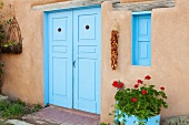 Einfache Hausfassade mit strahlend blauer Eingangstür; davor leuchtend rote Geranien und ein Chilizopf