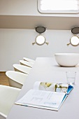 Open book and china bowl on table with white shell chairs in houseboat cabin
