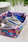 Patterned plastic container of cutlery in front of stacked plates and glasses on tablecloth in garden