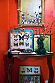 Red wall as backdrop for collection of framed butterflies and antique mirror with passport photographs stuck in frame