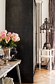 Silver, trophy-shaped vase of roses on wooden bench next to roller-door cupboard and iron lamp on side table