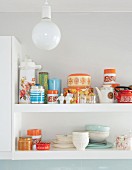 Detail of crockery and storage jars on white wall-mounted shelves behind spherical ceiling lamp