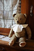 Old teddy bear wearing bib sitting on kitchen dresser in front of lace curtain in glass door