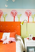 Colorful child's bedroom - orange upholstered headboard and bed linen in front of a tree wall stencil