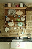 Various plates, cups and small jugs hanging from hooks on nostalgic kitchen shelving above vintage sink unit against stone wall