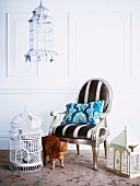 Brown tabby cat between an assortment of romantic birdcages and silver lacquered, upholstered baroque armchair
