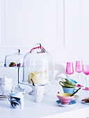 Pastel and antique style white porcelain, cake stands with glass covers and pink aperitif glasses and colorful cups on a white table
