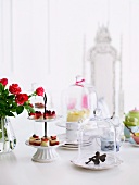 A table with a feminine touch - an elf under a glass cover, delicate cake stand and bouquet of roses. In the background a throne like white chari
