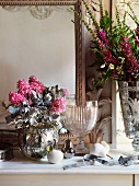 Blumensträusse in versilberten Vasen und Kristallschale mit Fuss auf Ablage vor Spiegel im eleganten Ambiente.