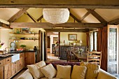 View from kitchen-dining area into foyer of simple English country house with exposed timber structure; comfortable sofa with many scatter cushions in foreground