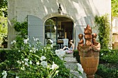 weiße Gartenblumen und Terrakottadekoration in sommerlichem Garten; im Hintergrund eine offene Terrassentür mit davor sitzender Frau