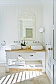 Solider Waschtisch aus Holz im Landhausstil mit darüberhängendem, gerahmten Spiegel und runden Waschschüsseln; dazwischen weiße Orchideen