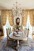 Magnificent dining room with solid wooden dining table below pretty chandelier; gathered, floor-length curtains in background at windows with view of park