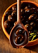 Schwarze Oliven in Olivenöl auf Holzlöffel und in Tonschale
