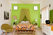 Bedroom with green mosquito net over double bed against green wall and flanked by green shell chair and white sideboard
