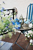 Silbertablett mit ausgefallenen, antiken Teekannen auf blau lackiertem Gartenstuhl; Terrasse mit floralem Brüstungsgeländer