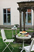 Afternoon tea in garden with antique bistro table and white vintage chairs in front of historical stone pavilion
