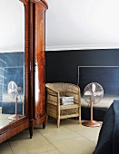 Großer, antiker Spiegelschrank mit edlem Holzfurnier; daneben ein einfacher Rattanstuhl und ein Ventilator