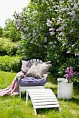 Gartenstuhl mit weichen Kissen und Tagesdecke unter prachtvoll blühendem Flieder; daneben eine weiss gestrichene Tonne als Beistelltisch