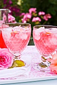 Two glasses of rose syrup with ice cubes and rose petals on a tray
