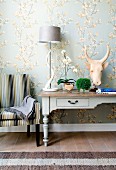 Antique country-house desk against wallpaper with delicate floral pattern contrasting with horned animal head sculpture and spiral horn as lamp base