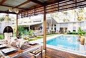 Dining table on roofed terrace with view of pool and luxurious, one-storey white house with pitch roof