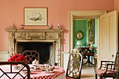 Pink breakfast room with rustic tablecloth on table in front of open fireplace with stone mantelpiece and view of adjoining room through open door