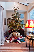 Traditionally decorated Christmas tree raised above mountain of presents in large bay window; lamp with glass balloon base and festive lampshade to one side