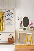 Dress and coathangers hanging on front of white-painted farmhouse wardrobe next to dressing table and yellow stool against white, wood-panelled wall