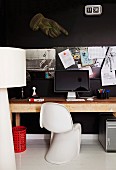 Classic shell chair in front of computer on wooden table against black wall with pinboard