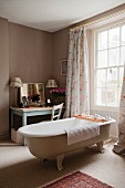 Freistehende Vintage Badewanne vor Fenster mit bodenlangem Vorhang, gegenüber Tisch mit Spiegel in Zimmerecke